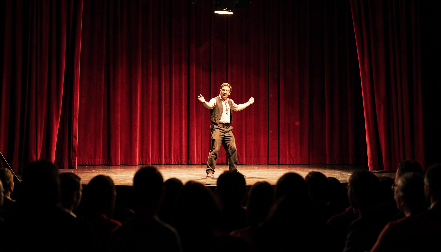 A warmly lit stage at a magic convention, deep red curtains framing a performer mid-routine, audience silhouettes in the foreground