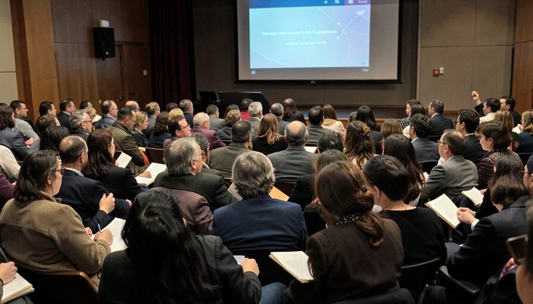 Rows of seated attendees at a magic lecture, notebooks open, projector screen visible